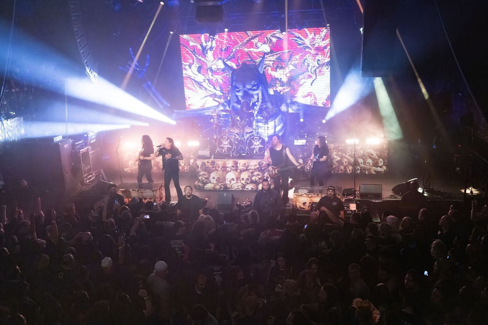 Testament live in concert. The band stands on stage performing in front of a large crowd in a packed theater room