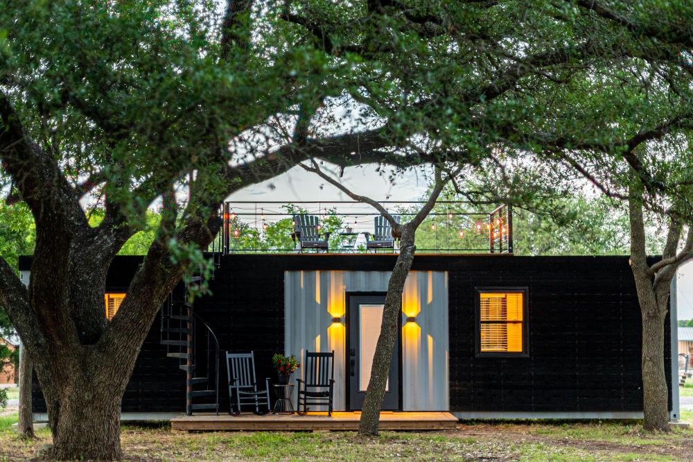A tiny house made from a conex box with lawnchairs, trees, and front yard.