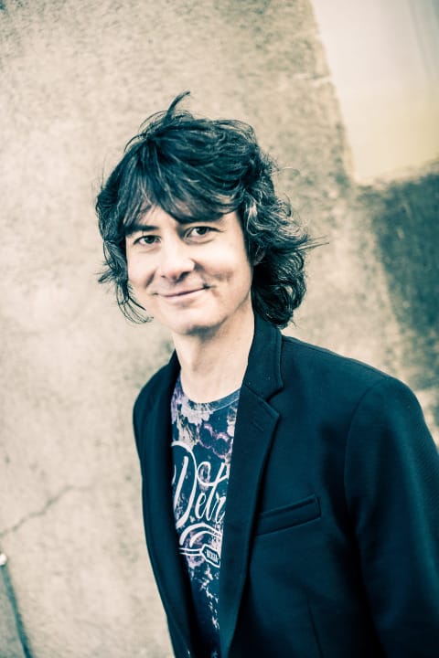 Author Antti Tuomainen wearing a blue blazer and T-shrit and smiling at the camera in a roguish way