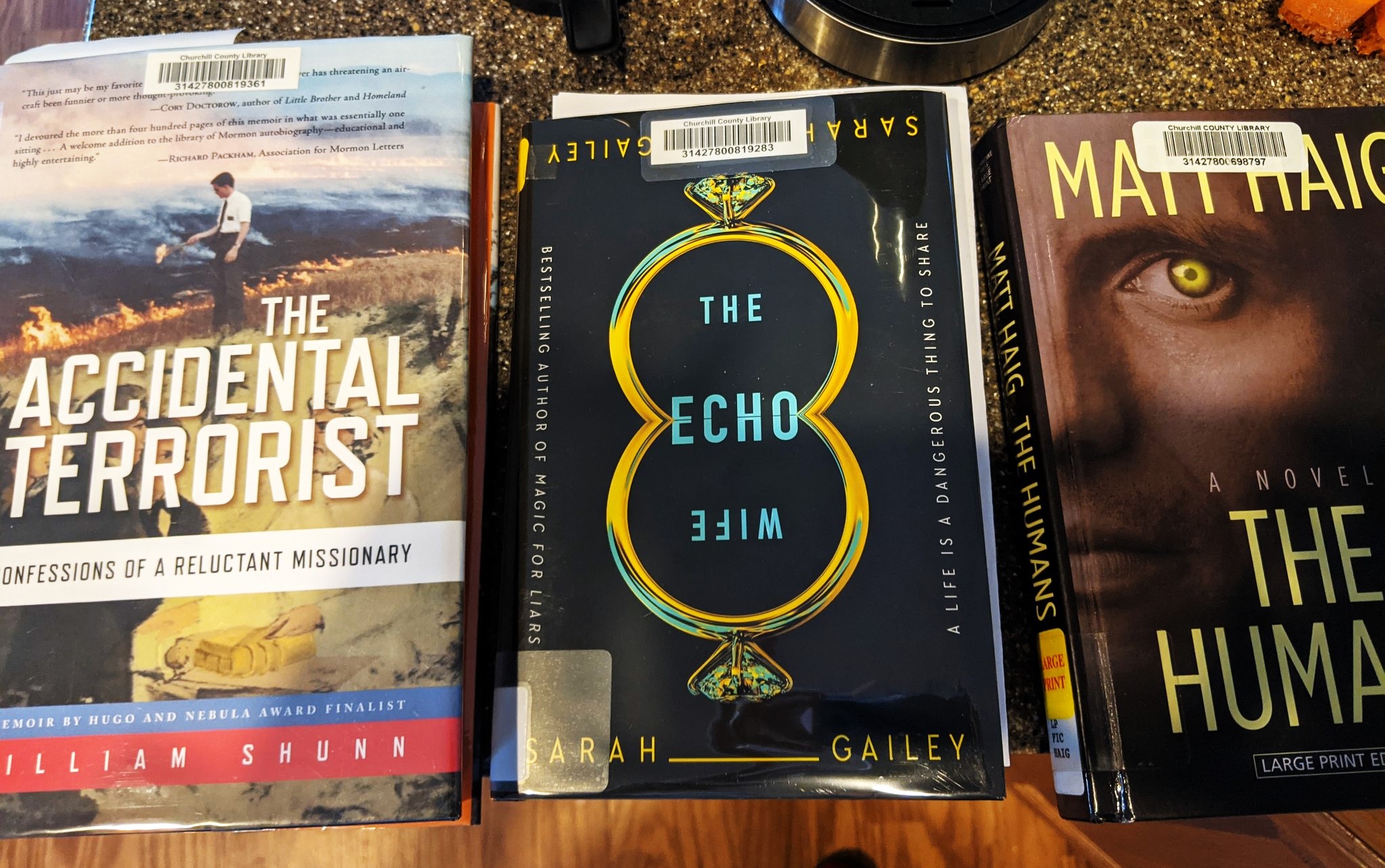 Three books (The Accidental Terrorist by William Shunn, The Echo Wife by Sarah Gailey, The Humans by Matt Haig) sit face-up side by side on a counter, all featuring bar codes from the Churchill Country Library in Nevada.