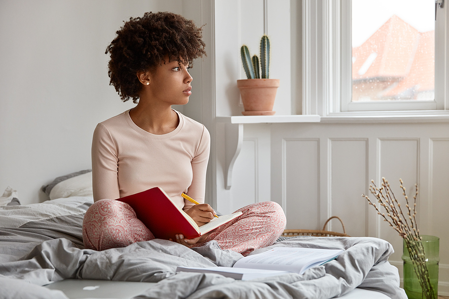 A thoughtful young woman sits on a rumpled bed in a bright bedroom writing in a notebook.