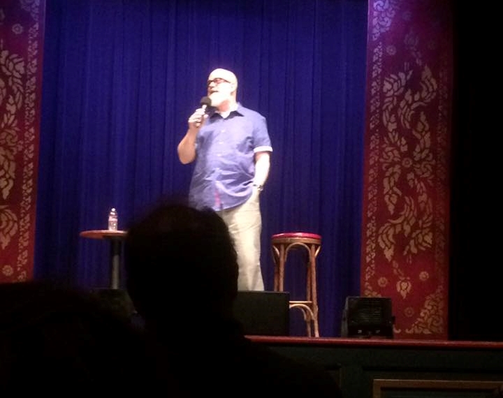 A bearded man stands on stage near a stool in front of a blue curtain in a classic-style theater, with the silhouetted heads of audience members in the foreground.