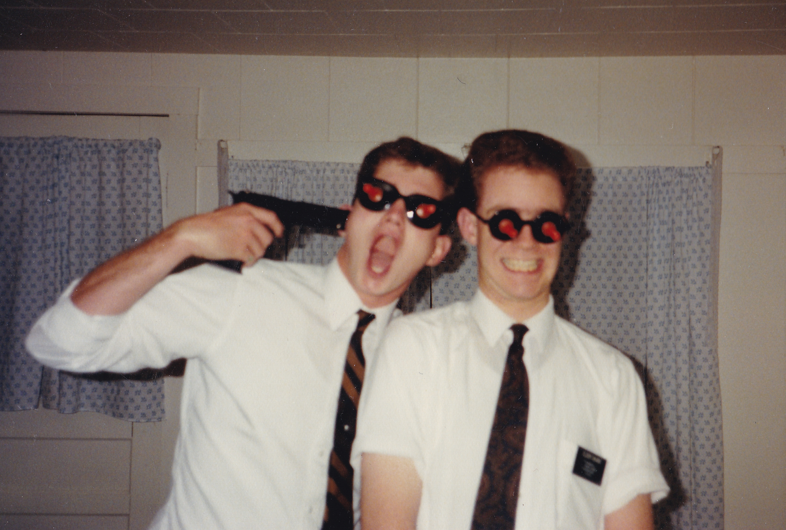 Two stupid young LDS missionaries stand side by side wearing dark sunglasses, with an orange rubber suction dart stuck to each lens. The missionary on the right is baring his teeth in a Joker-like grin. The one on the left, open-mouthed, is pointing a handgun at his own temple -- with the implication that one shot could take them both out.