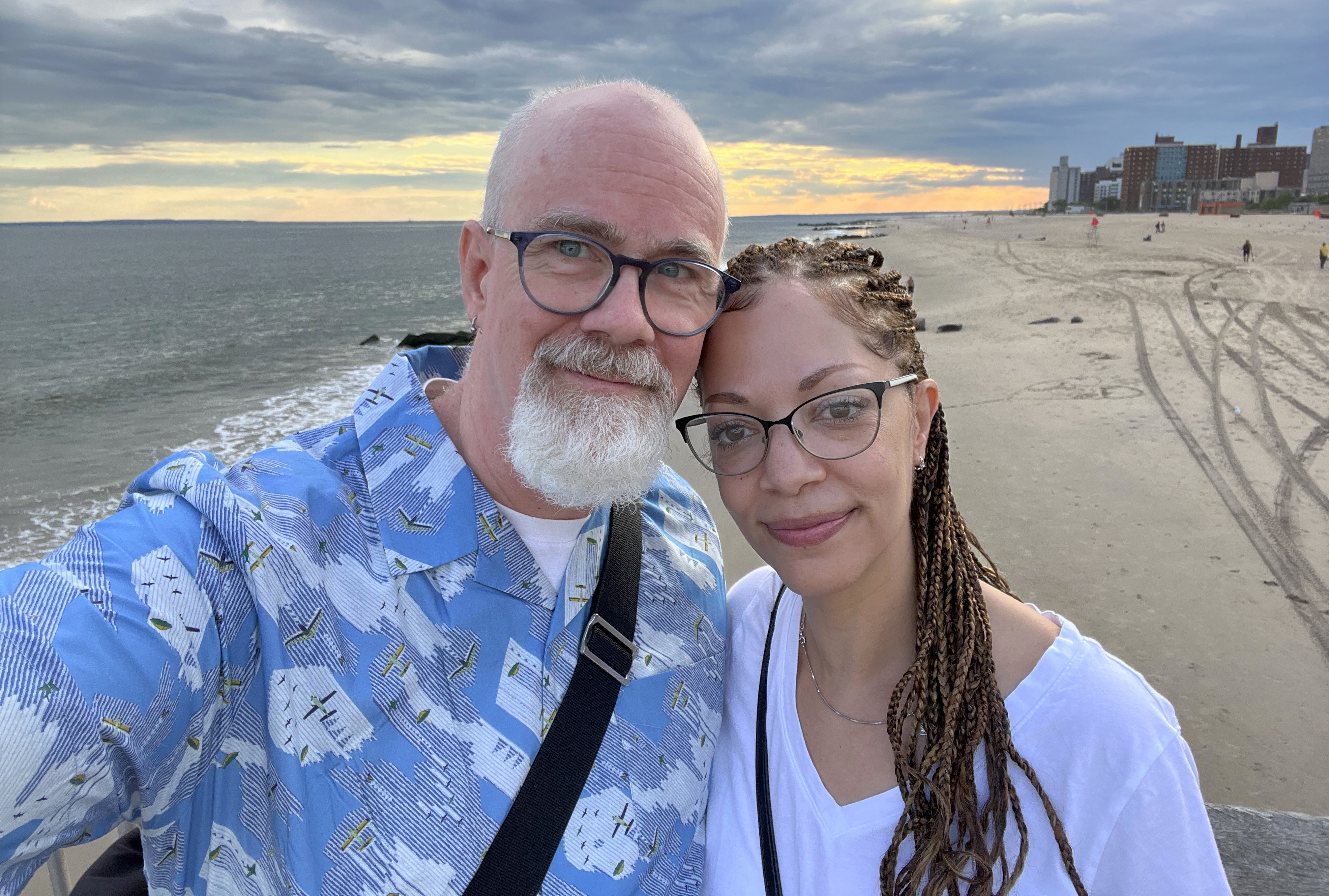 A man and woman lean together taking a selfie on a cloudy beach near dusk. He is bald with a gray goatee. She has long braids pulled back and hanging over her shoulder.