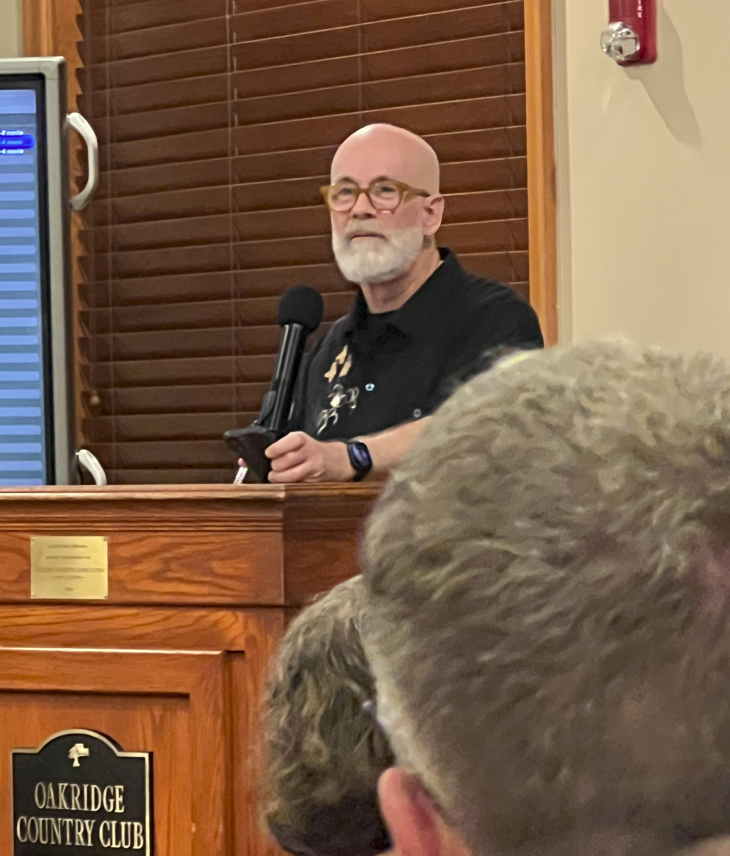 A somewhat blurred long-distance photo of William Shunn (a bald, bearded man in a black shirt) speaks at a lectern that reads "Oakridge Country Club."