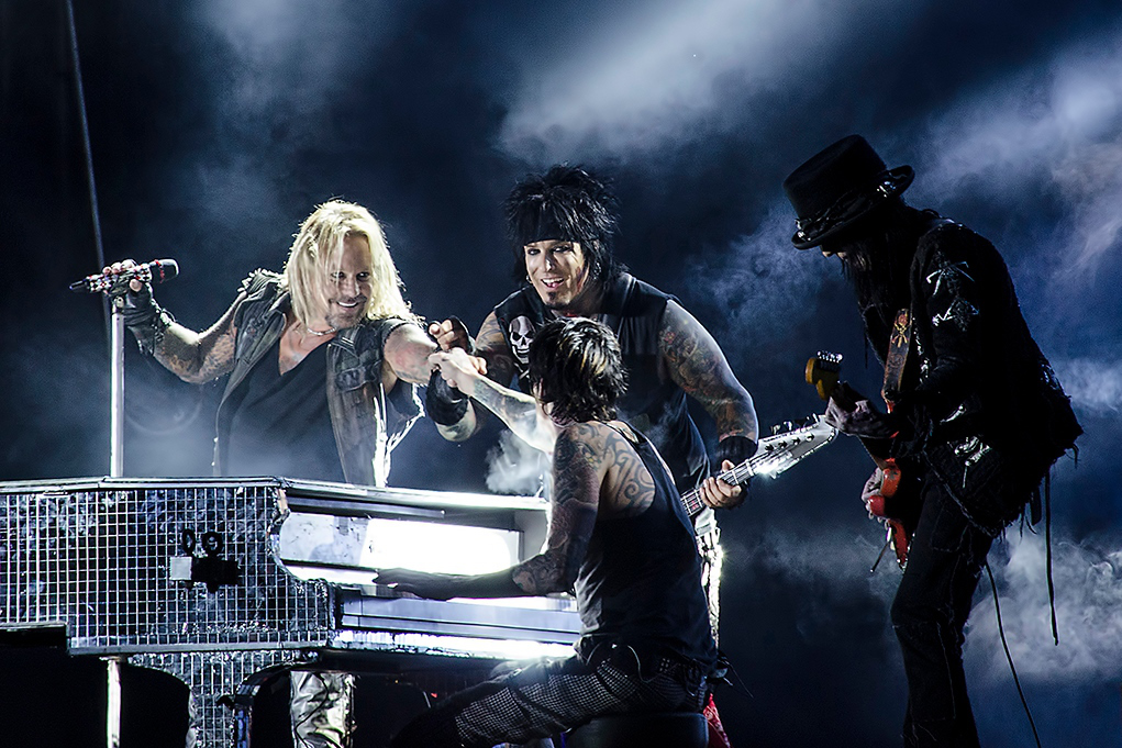 A hair metal band on stage, gathered around a piano covered with mirror tiles. The drummer is, for some reason, seated at the piano, and he is giving the lead singer a fist dump.