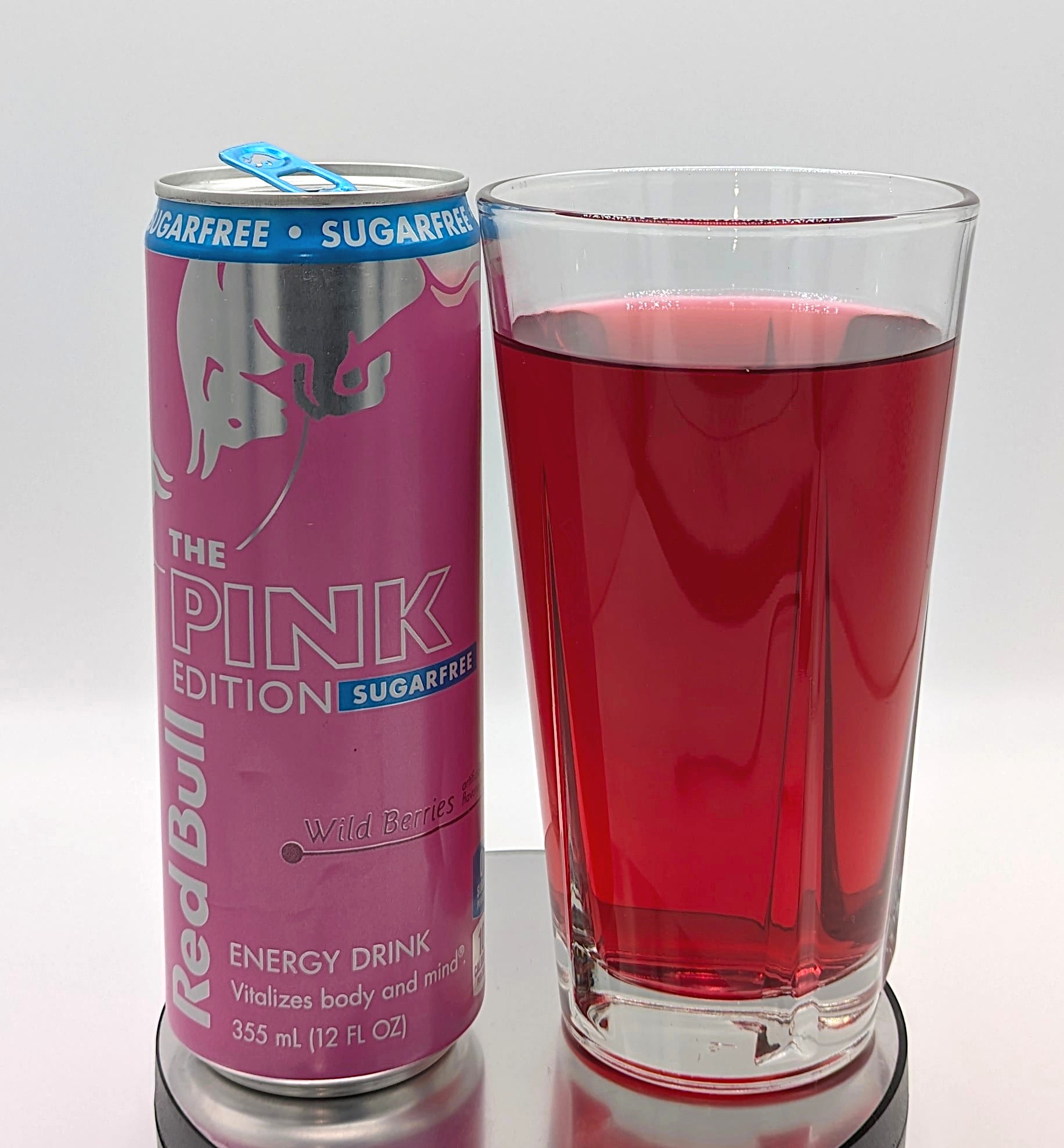 A can of Red Bull Pink Edition Sugar Free next to a glass containing the drink. The liquid is a wine red.