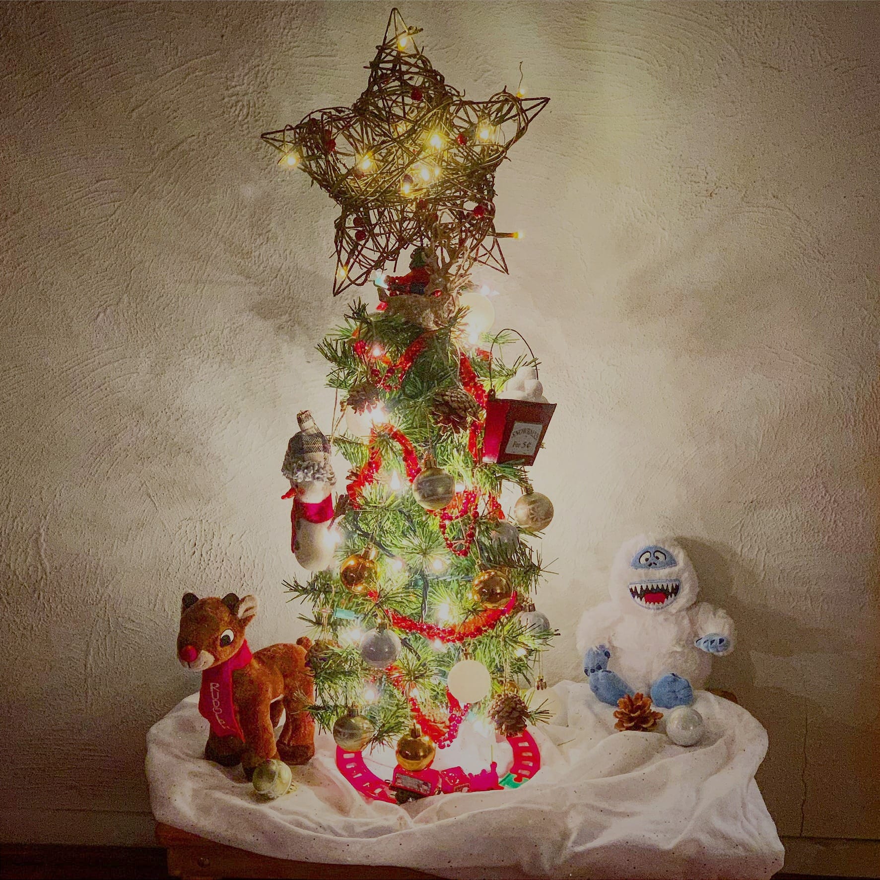 A small lit Christmas tree on a table draped in white cloth, against a pale textured wall. A twig star with tiny lights tops the tree, which is hung with red garland and silver and gold ornaments. A plush reindeer in a red scarf stands at left, a plush white snowman monster at right, and a small pink toy train track circles the base.