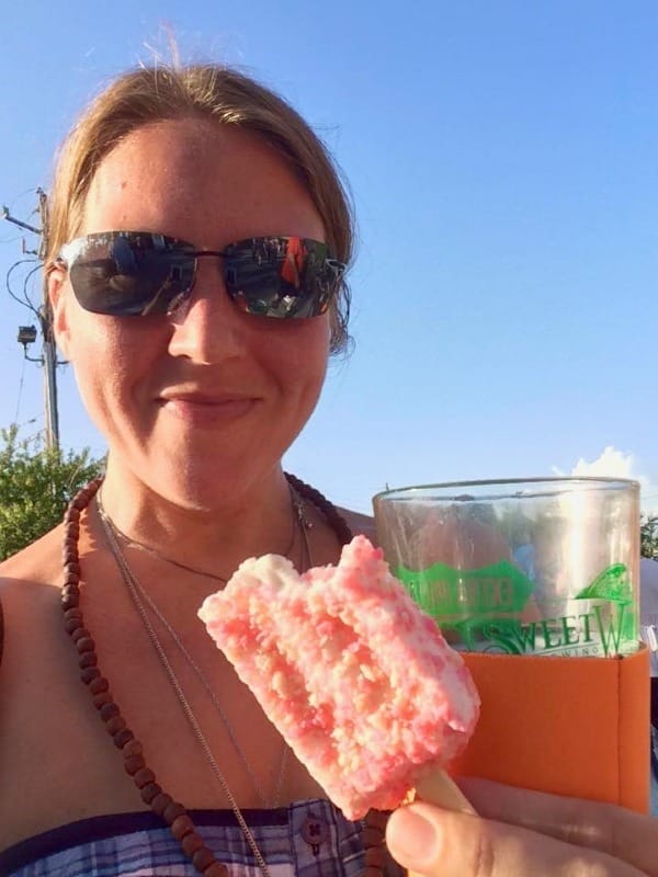 The author outdoors on a bright day in sunglasses, smiling slightly, holding a pink ice cream bar toward the camera with a brewery cup in her other hand.