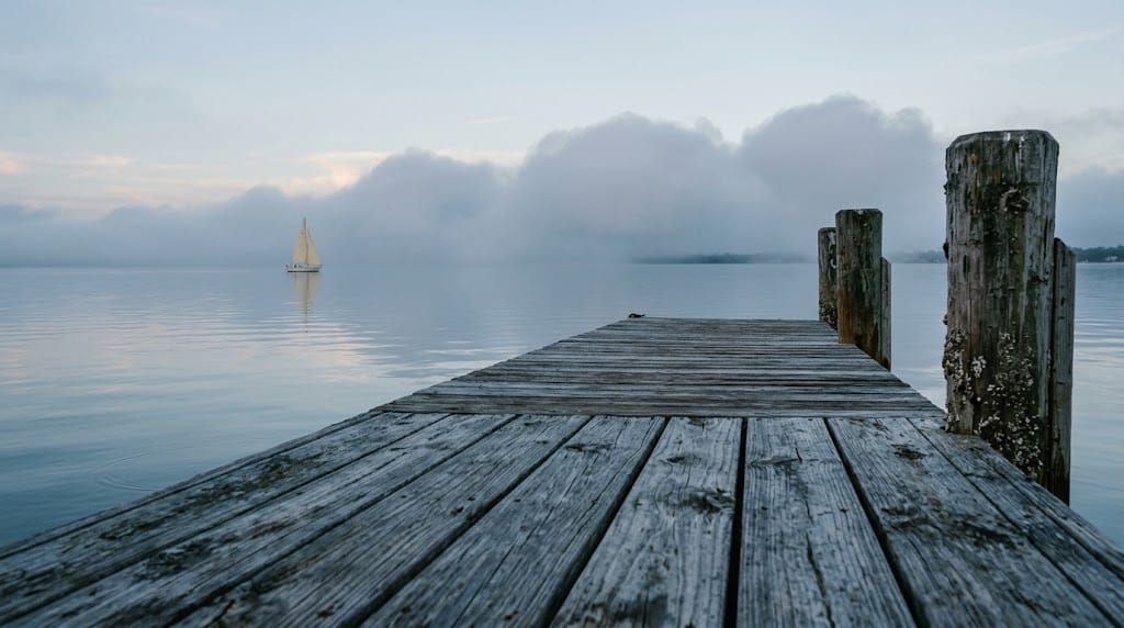 A weathered wooden pier with barnacled pilings reaches over still water in early light; far off on the left, a lone small sailboat sits beneath a low bank of fog.