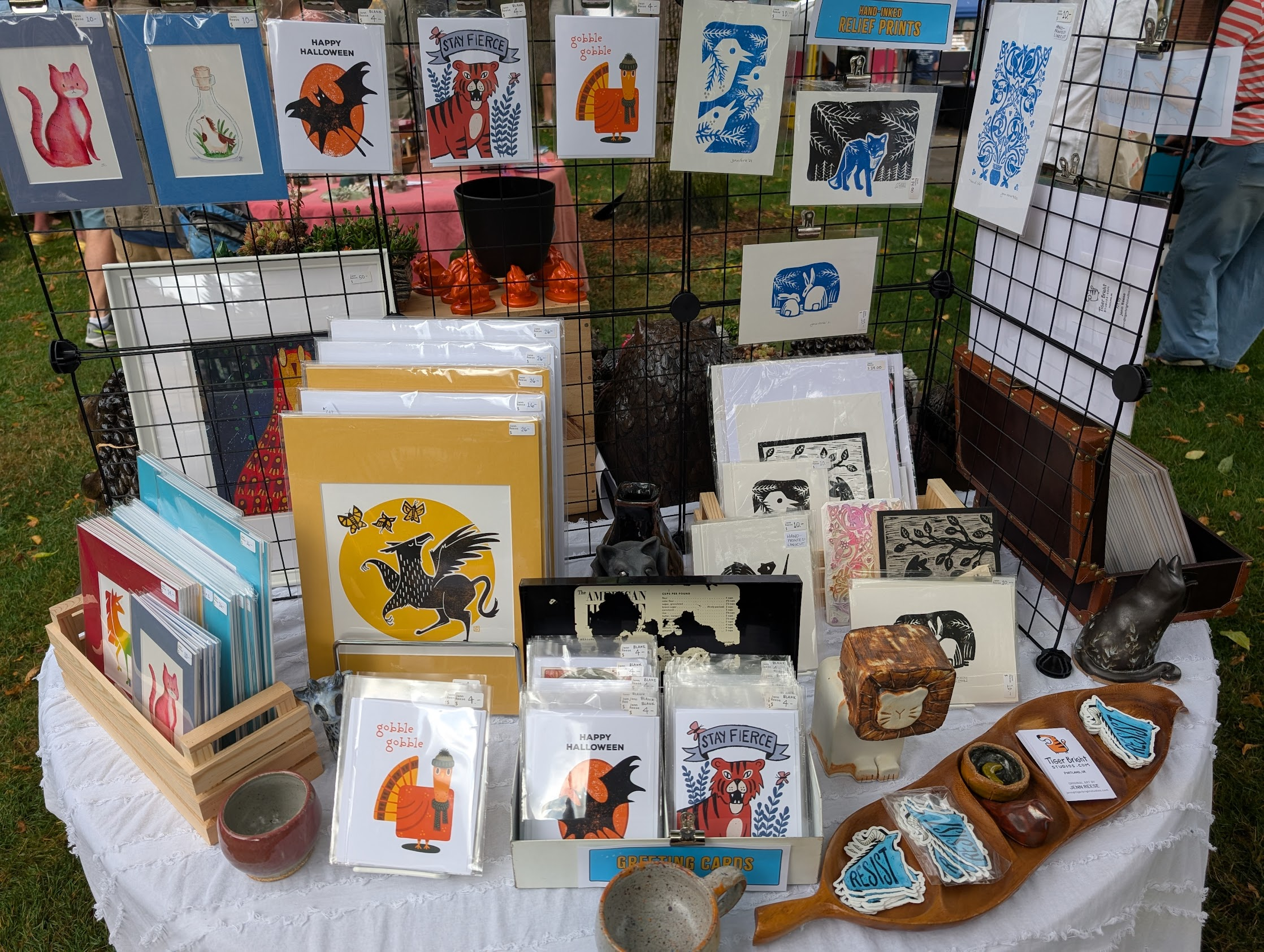 Half of a circular table jam-packed with stacks of art prints in bright mats, cards hanging from wire shelves, and a tray with stickers.