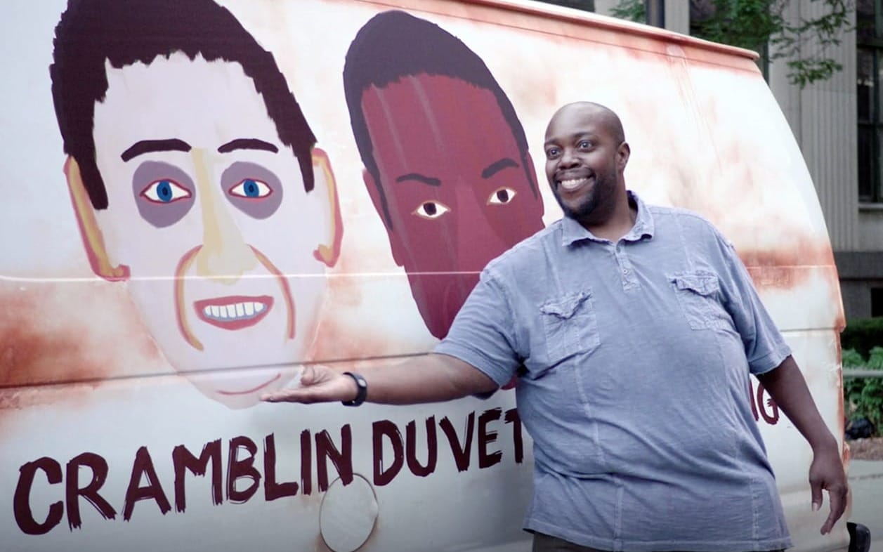 A screenshot from the show The Detroiters. A man motions to a terrible painting of two faces on the side of a van.