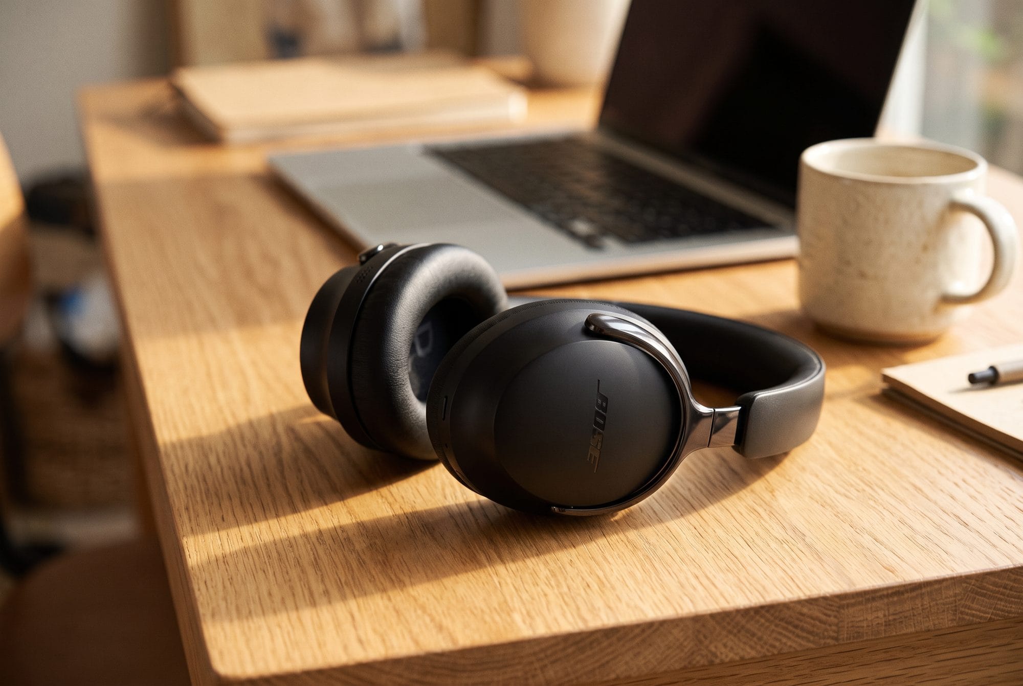 AI-generated image of black Bose QuietComfort Ultra headphones (2nd Gen) resting on a wooden desk beside a laptop and a cream mug in warm afternoon light.