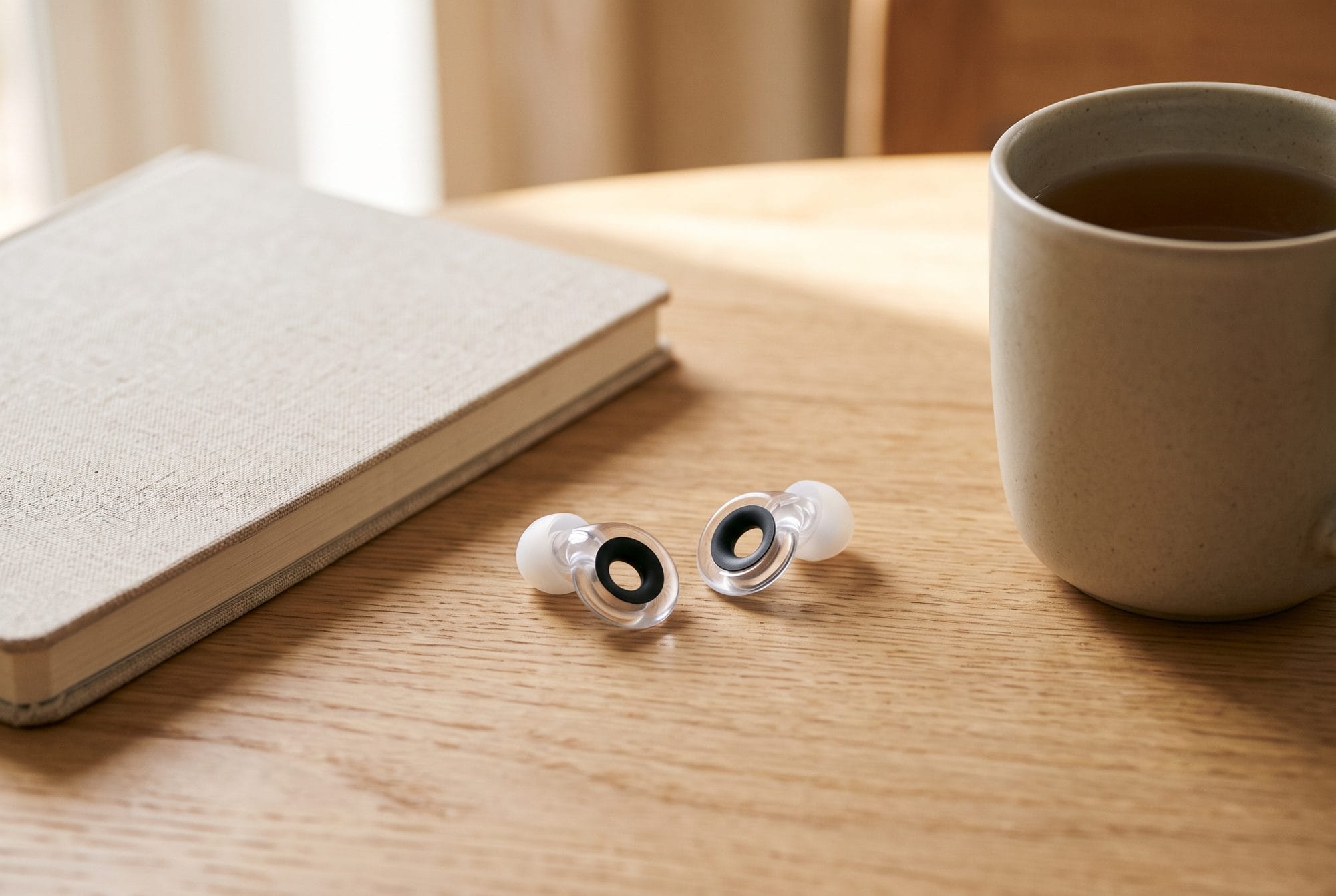 AI-generated close-up of a pair of clear Loop Engage 2 Plus earplugs with black Mute inserts on a wooden table beside a notebook and a mug of tea.