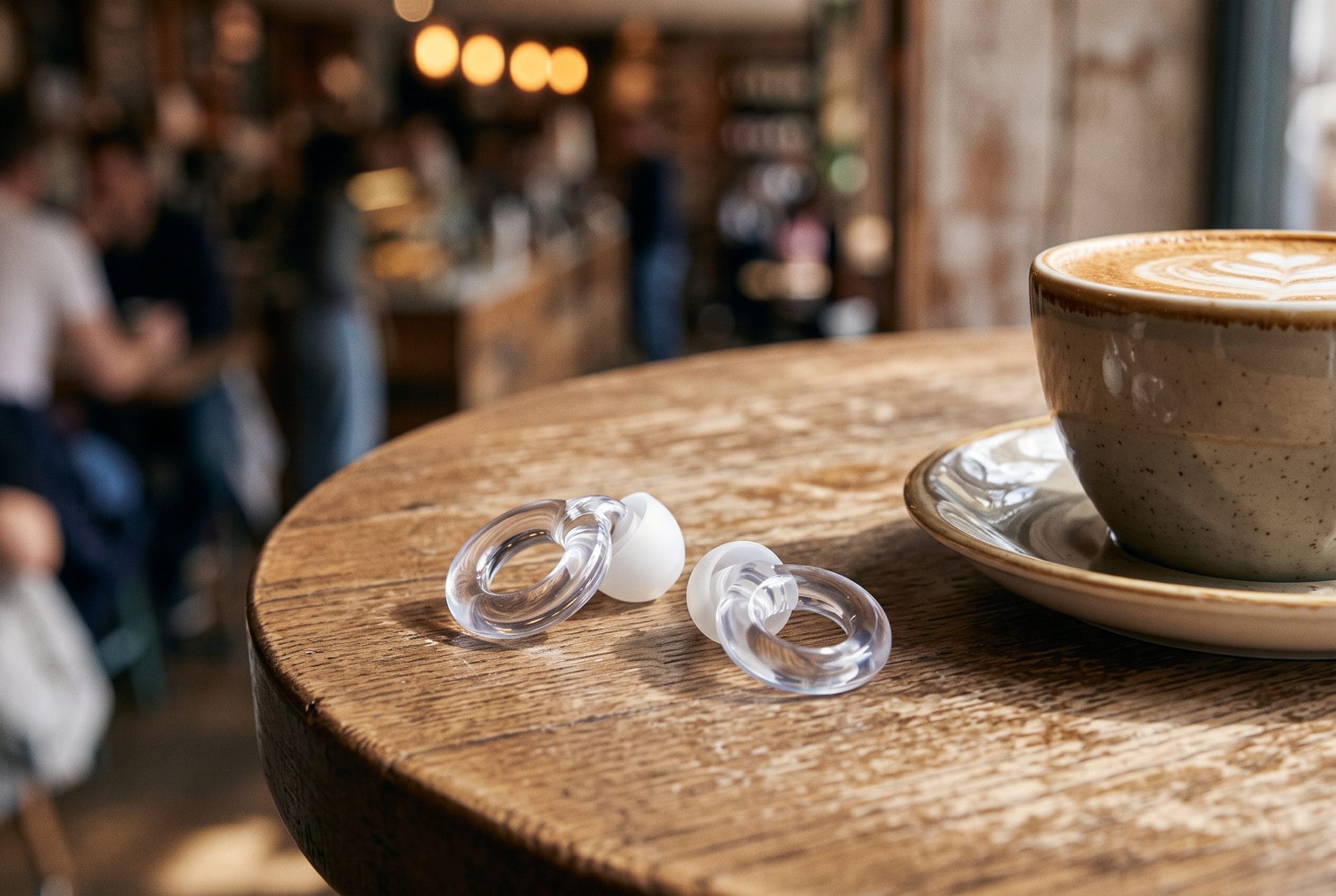 AI-generated close-up of a pair of clear Loop Engage 2 earplugs on a wooden café table beside a cappuccino, with a busy café blurred behind.