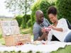 A heterosexual couple lay on a picnic blanket in a park. The woman is peeling open a plastic tray full of raspberries. An…