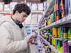 A man wearing headphones, stood in a supermarket isle, looking at a can of energy drink