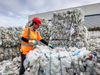 A worker wearing a safety helmet and hi-vis jacket stands amongst towering piles of plastic bales. She is reaching out to…
