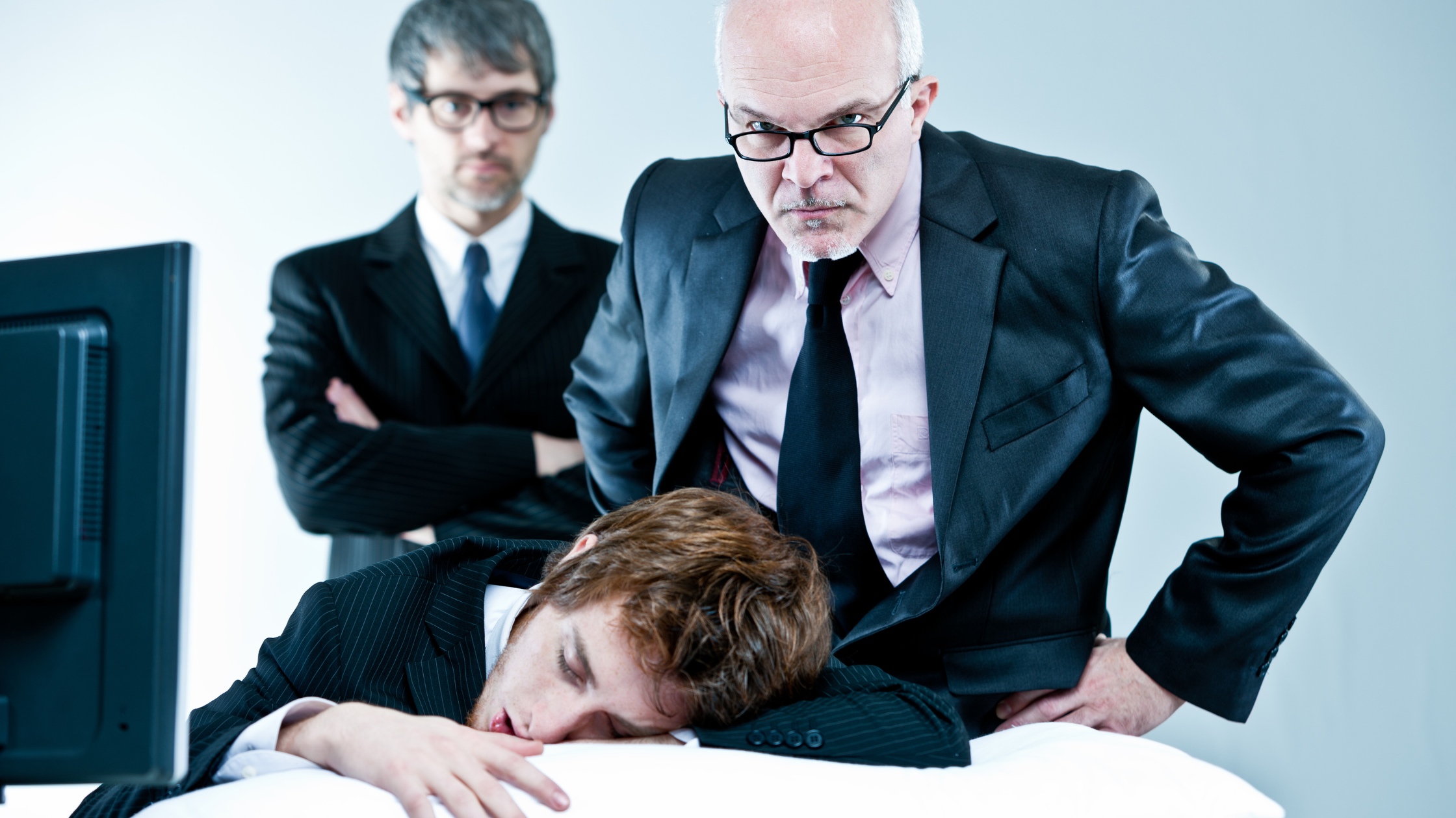 Young office worker asleep at desk while two stern managers in suits look on disapprovingly