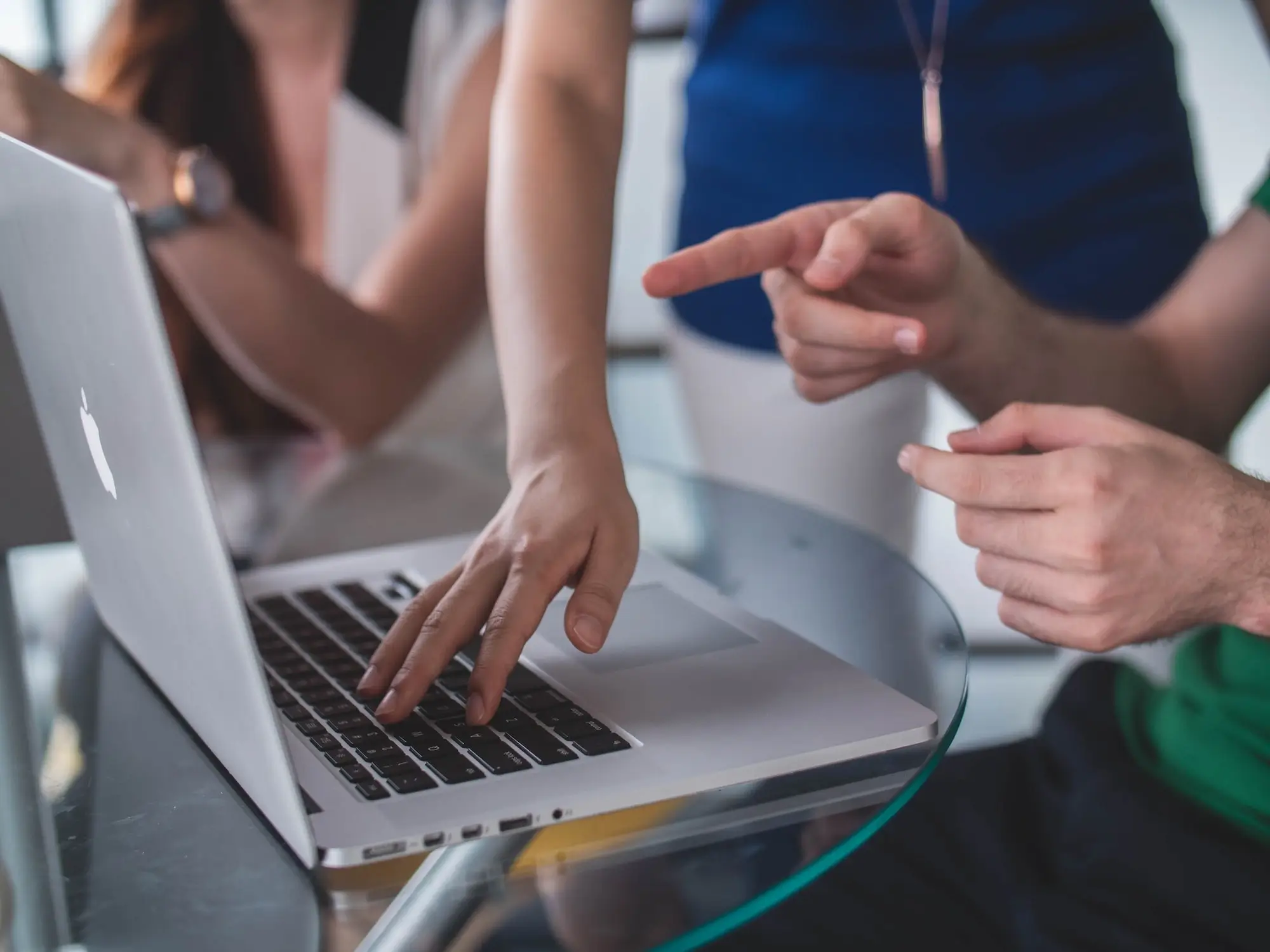 Drei Softwareentwickler sitzen an einem runden Glastisch, vor ihnen ein Macbook, eine Hand ist auf der Tastatur, die anderen beiden erklären und zeigen auf den Bildschirm