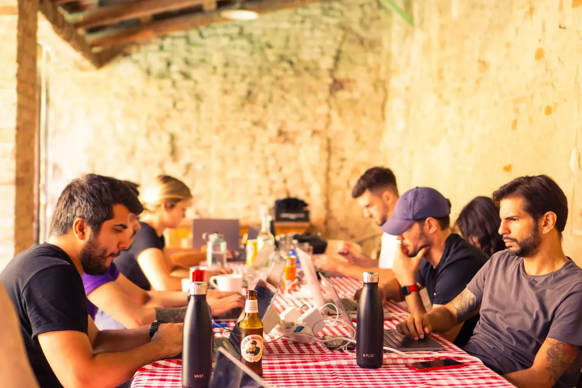 Eine Gruppe Quality Assurance Manager arbeitet an einem Tisch im Freien mit Macbooks, im Hintergrund eine Backstein-Wand. 
