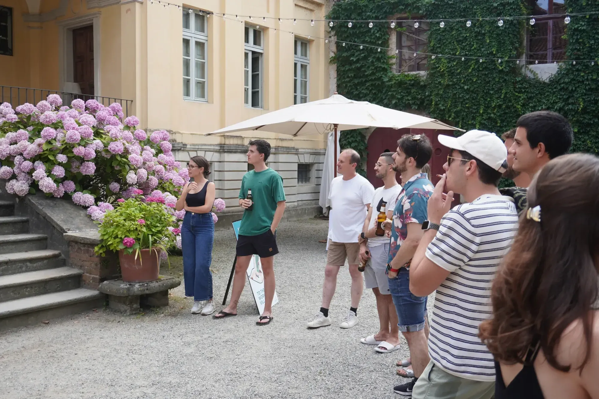 Eine Gruppe von Menschen steht in einem Innenhof und blickt nach links, in der Hand Getränke. Ein großer Blumenstrauch auf der linken Seite, ein gelbes Gebäude in der Mitte, ein beiger Sonnenschirm und eine mit Efeu überwucherte Hausfassade auf der rechten Seite. 