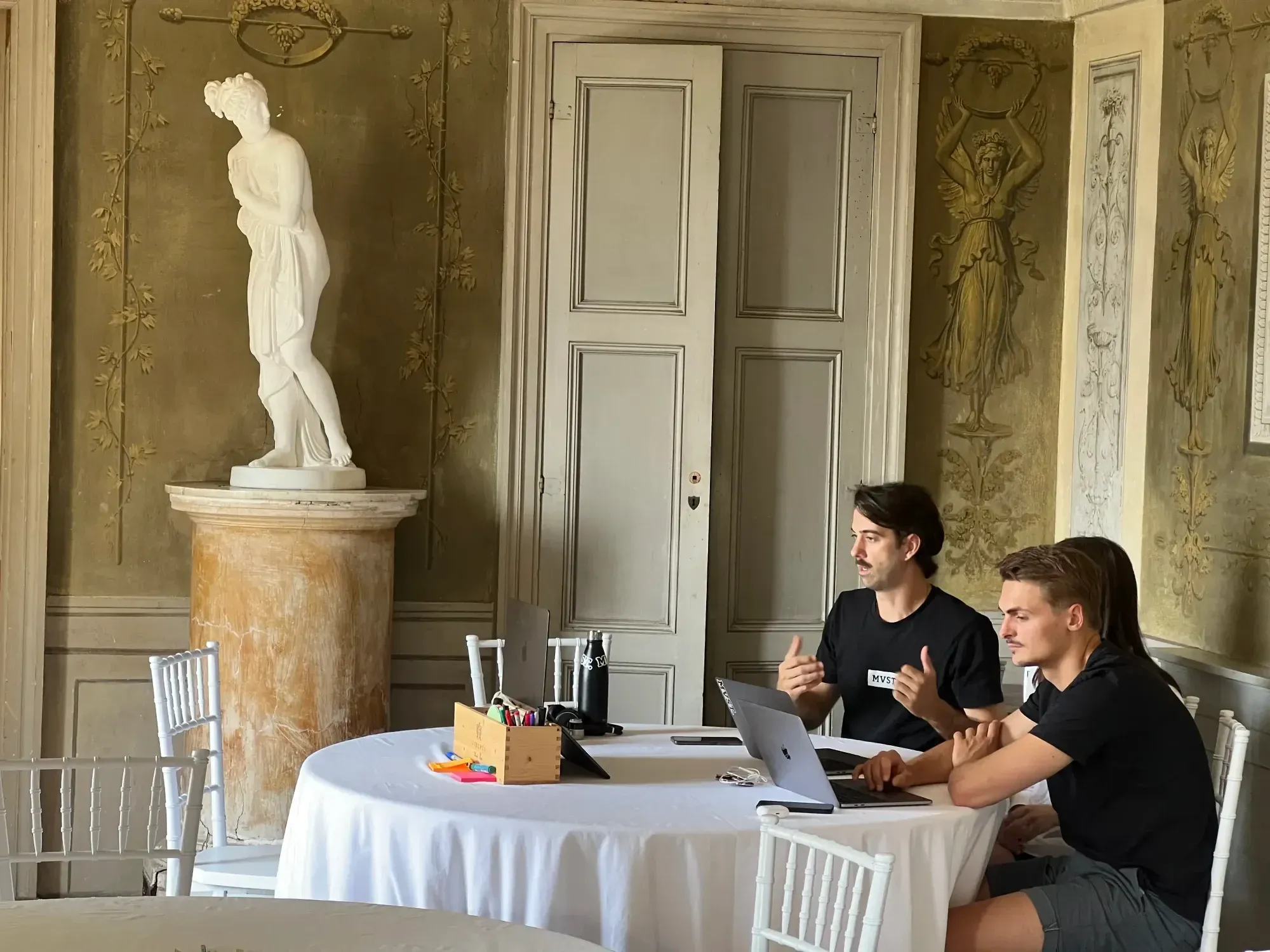 Drei Personen sitzen an einem runden weißen Tisch und arbeiten an ihren Macbooks. Der Raum ist mit einer großen weißen Statue einer Frau dekoriert und die Wände sind mit Ornamenten und antiken Zeichnungen bemalt.