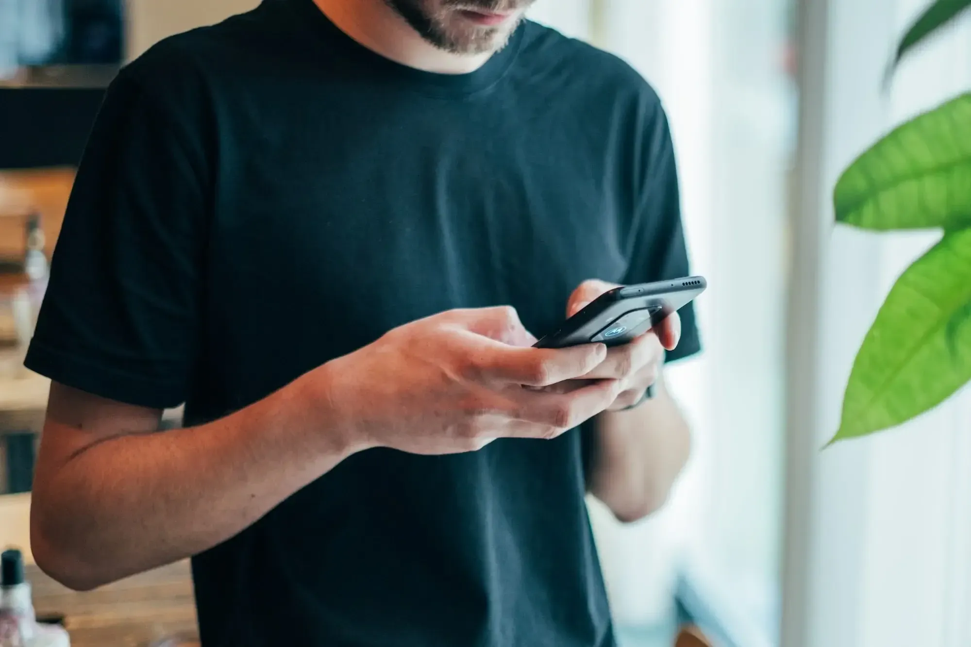 Guy with a black shirt holding a black phone with his hands, looking at it, plant next to him.
