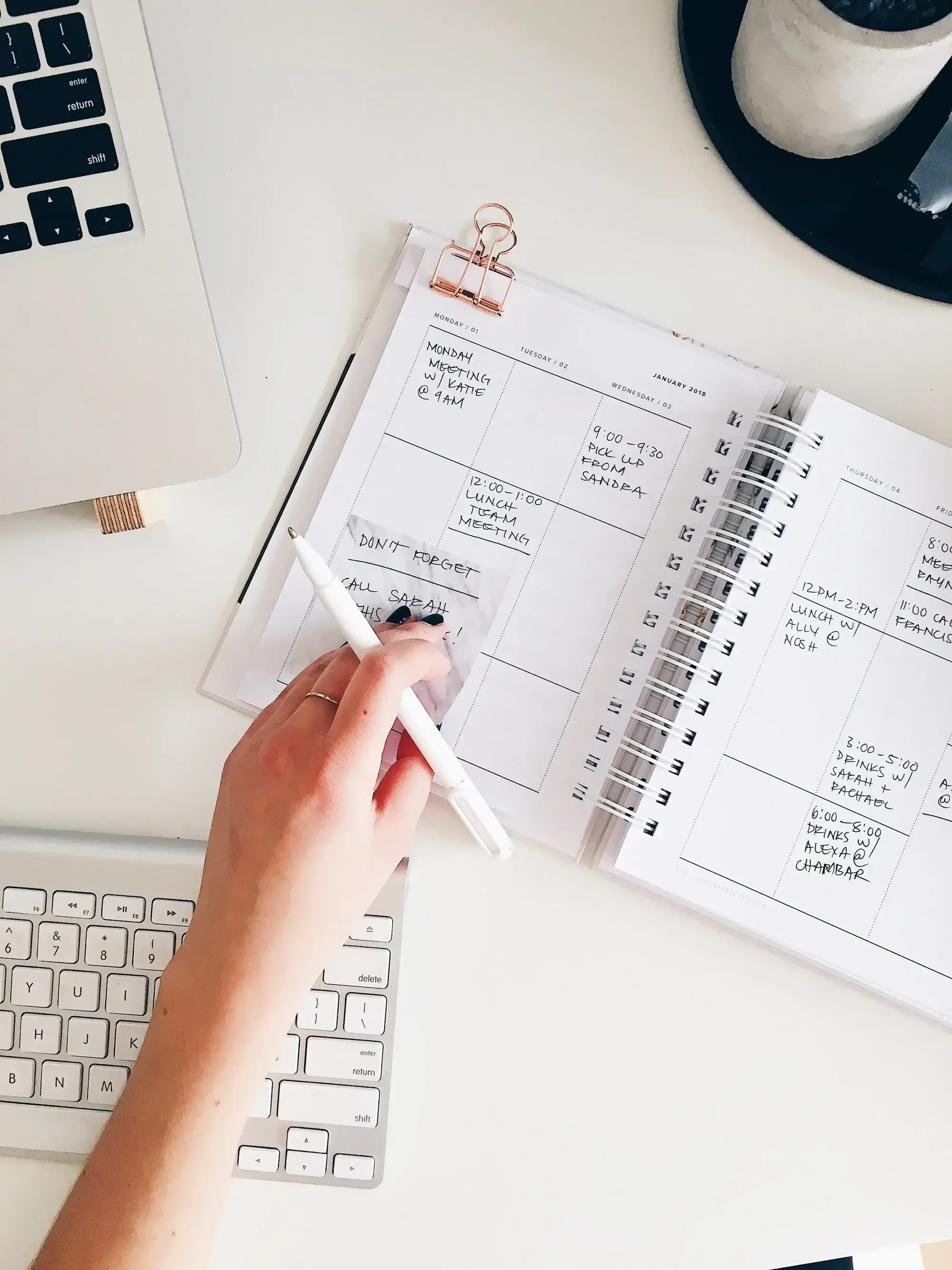 Hand of a woman holding a white pen, writing notes into her notebook, in the background a mac keyboard