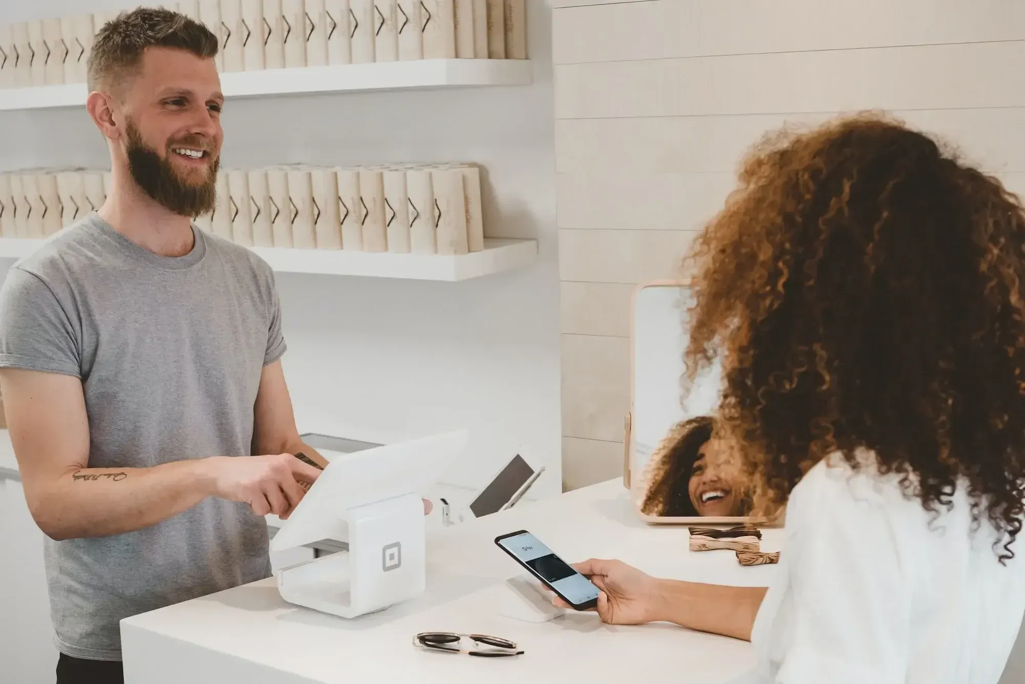a bearded young man with a grey shirt serviced a woman with curly hair and a white blouse as she pays with her phone