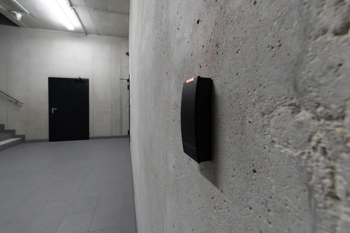 Black proximity card reader mounted on a concrete wall beside a secure door, the entry point access control records log.