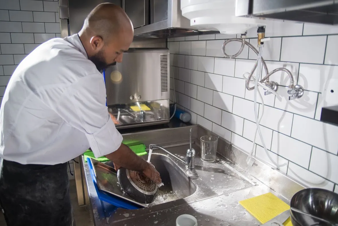 A chef in whites washing a pan in a stainless steel sink, the daily cleaning task a food business logbook records.