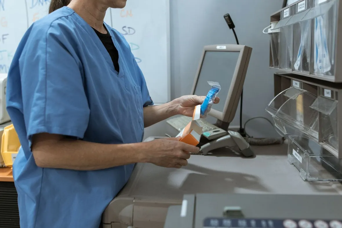 A healthcare worker in blue scrubs opening a carton at a medicine station with labelled storage bins, where controlled drug entries are recorded.