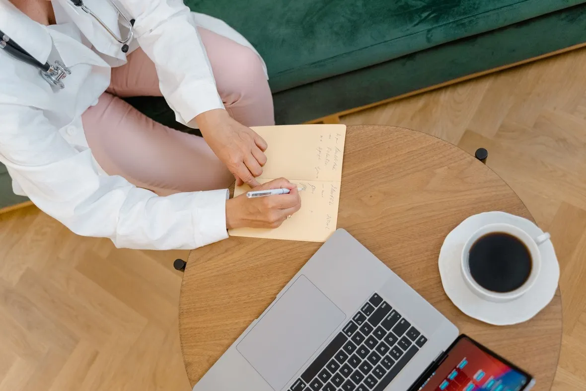A clinician with a stethoscope writing notes in a notebook beside a laptop, logging CPD hours for UK revalidation.