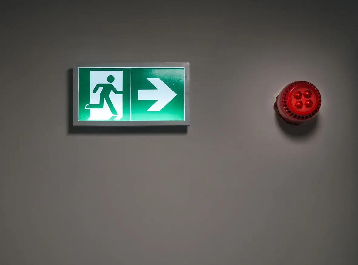 An illuminated green running man exit sign beside a red emergency beacon on a plain wall, the fittings a monthly logbook test covers.