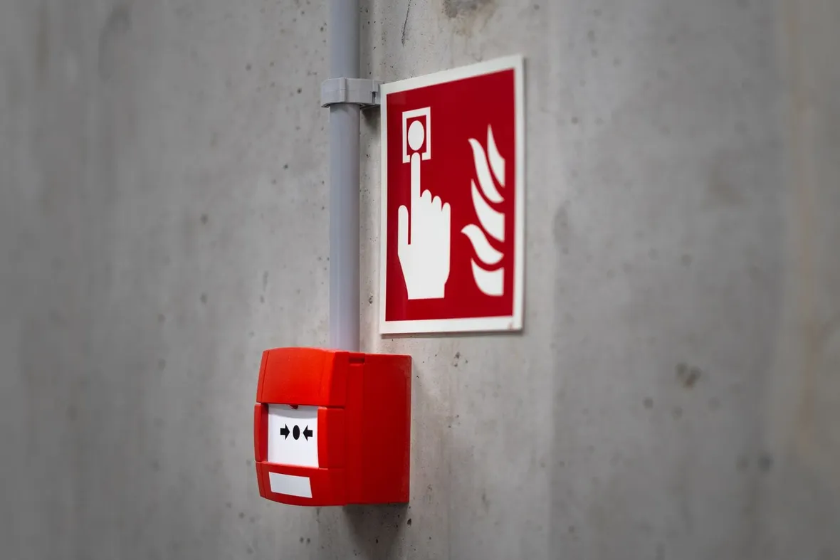 A red manual fire alarm call point beside a red pictogram sign on a bare concrete wall, the equipment a fire alarm logbook records.