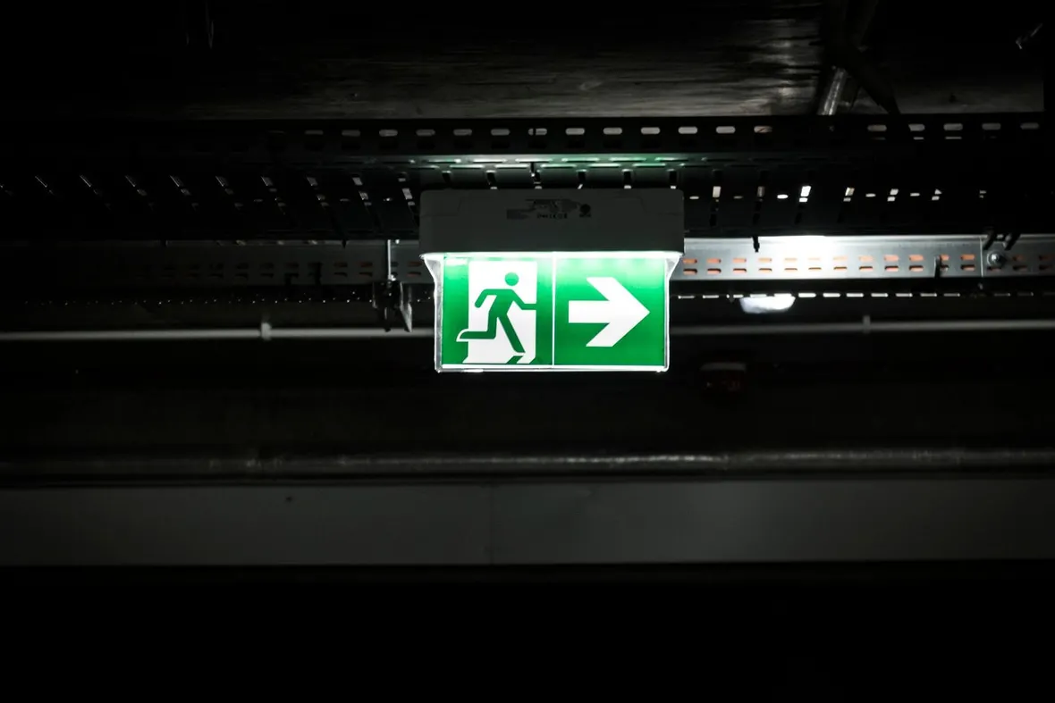 An illuminated green running man sign with an arrow below an industrial ceiling tray, marking the escape route a fire risk assessment must record.