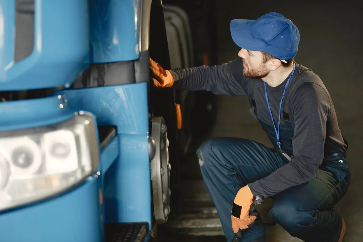 A worker in overalls crouching with a torch to inspect a lorry's front wheel, the daily walkaround check fleets must record.