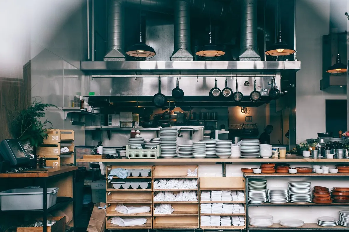 A commercial kitchen pass with stacked plates, hanging pans and overhead extraction ducting, the setting inspected for food hygiene record keeping.