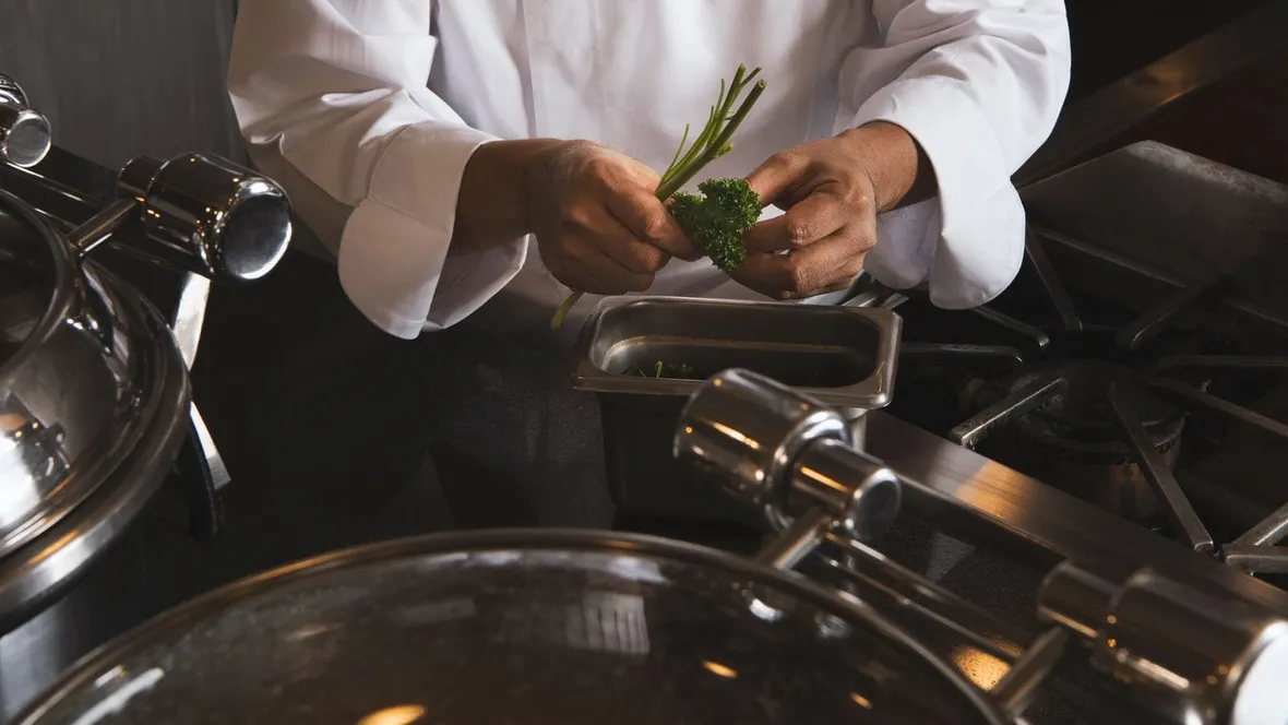 Chef's hands in white uniform preparing herbs into a stainless steel pan in a commercial kitchen, where food safety logbooks are a legal requirement.