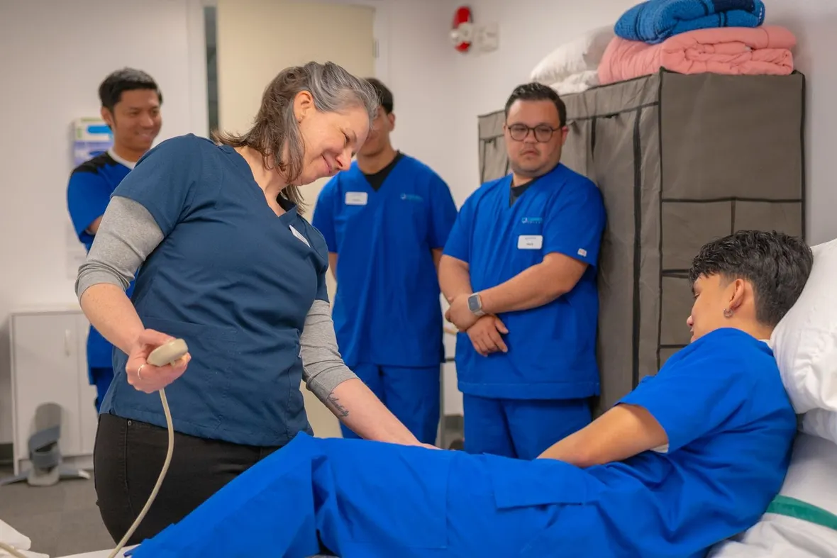 A clinical tutor demonstrating a bedside skill to trainees in scrubs, the supervised practice a healthcare training logbook records.