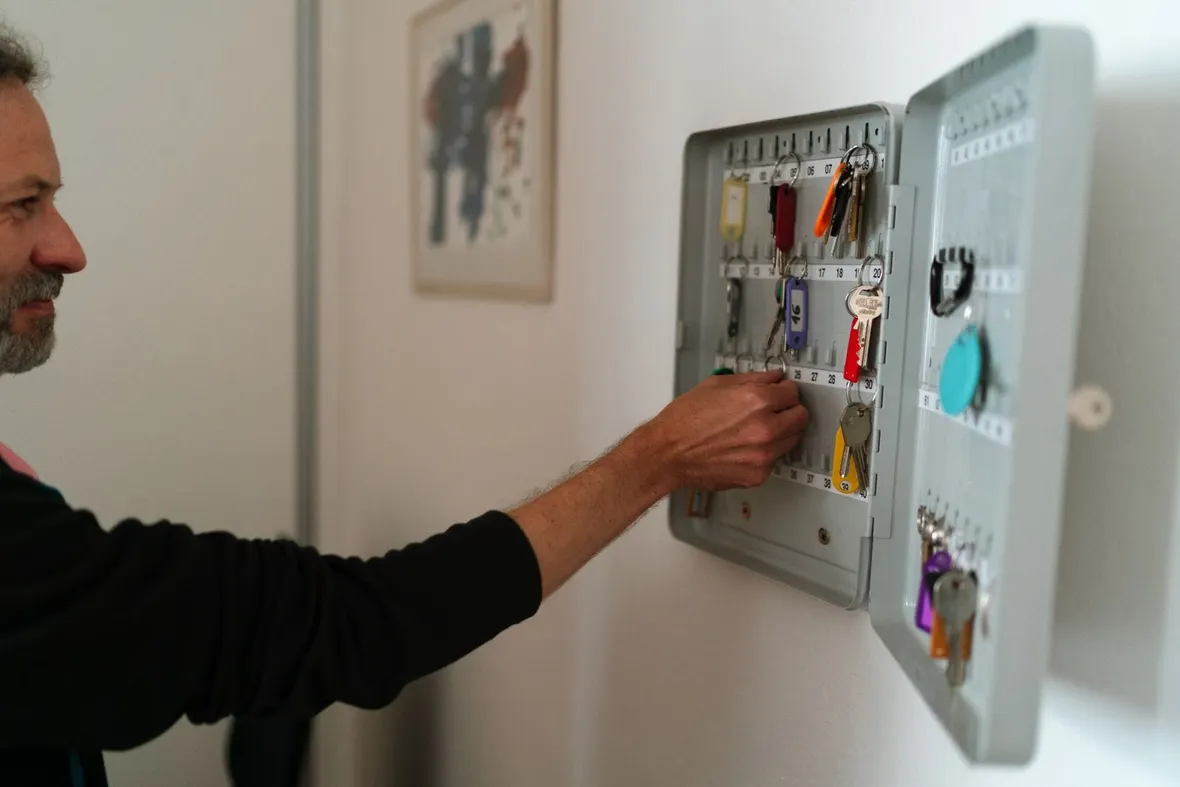 A hand reaching into a wall mounted key cabinet with numbered hooks and labelled keys, the basis of a key register.