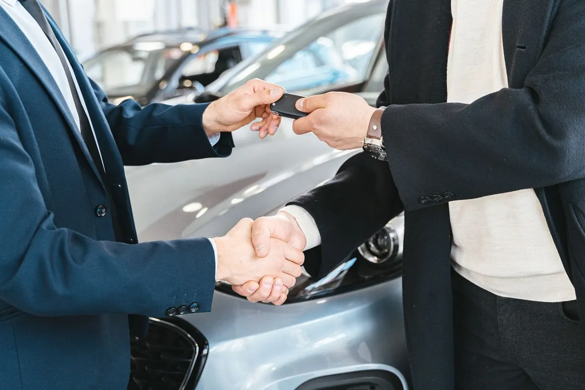 Two people shaking hands and passing a car key fob in front of a silver car, the kind of agreement behind a logbook loan.
