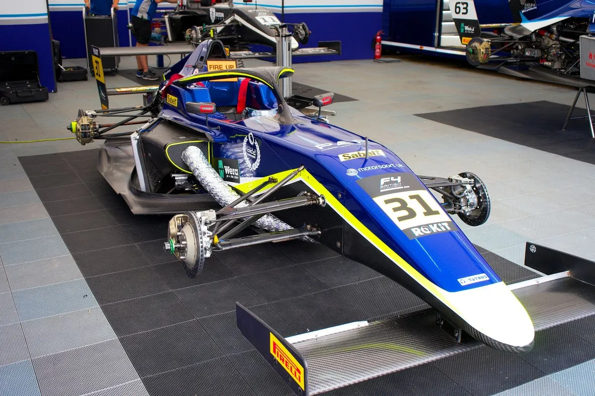 A British F4 single-seater with its wheels off and suspension exposed on garage matting, the kind of car scrutineers check before every event.