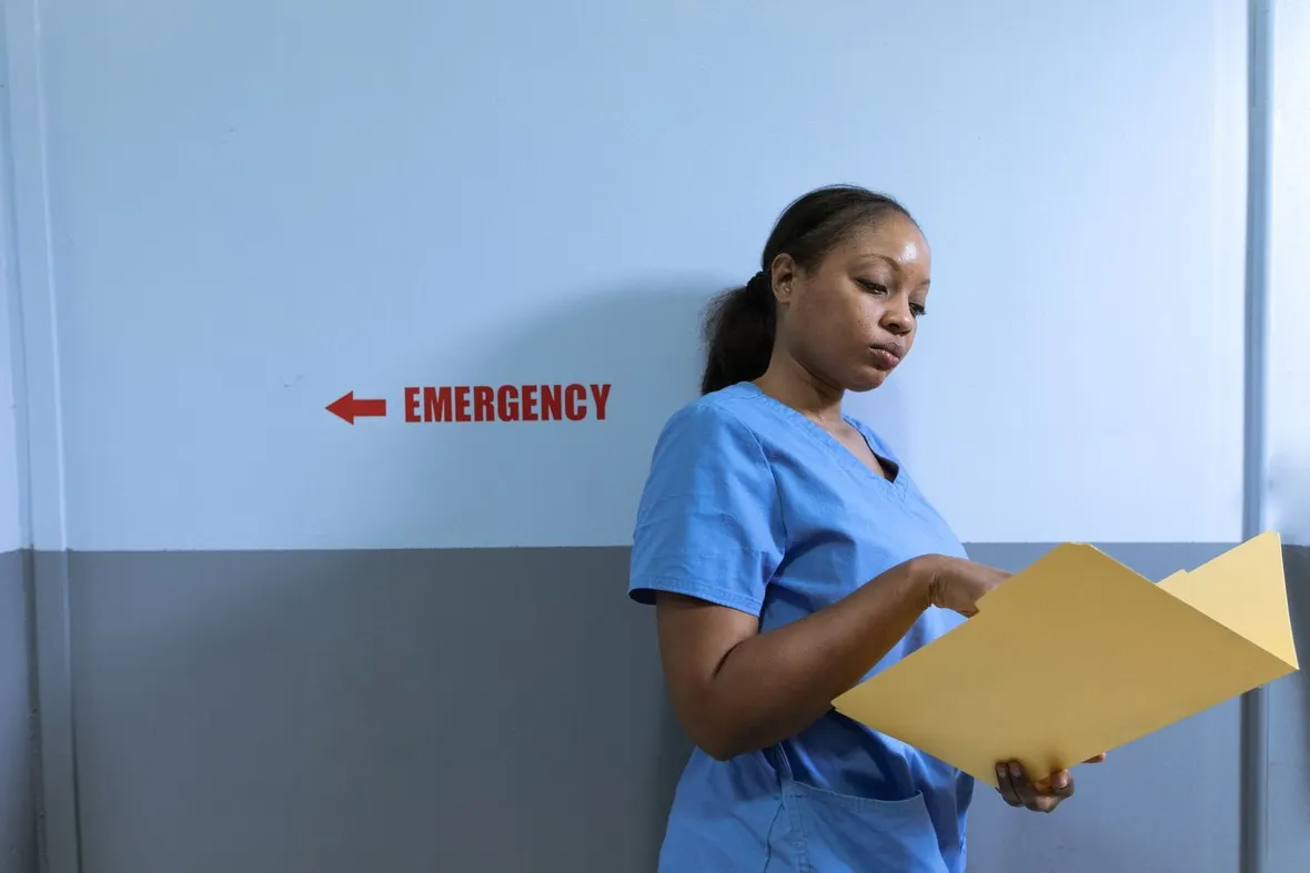 A nurse in blue scrubs studying an open card folder in a corridor beside a red emergency sign, as nursing logbooks record practice hours.