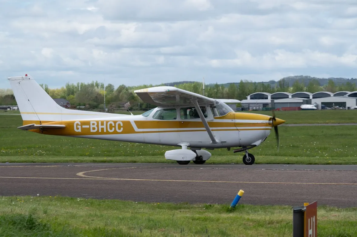 A Cessna 172 registered G-BHCC taxiing past grass and hangars at a UK airfield, where PPL training hours are logged.