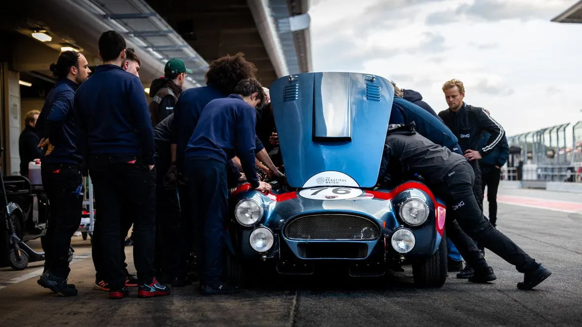 Mechanics crowded around a classic race car with its bonnet up in a circuit pit lane, where logbooks are checked.