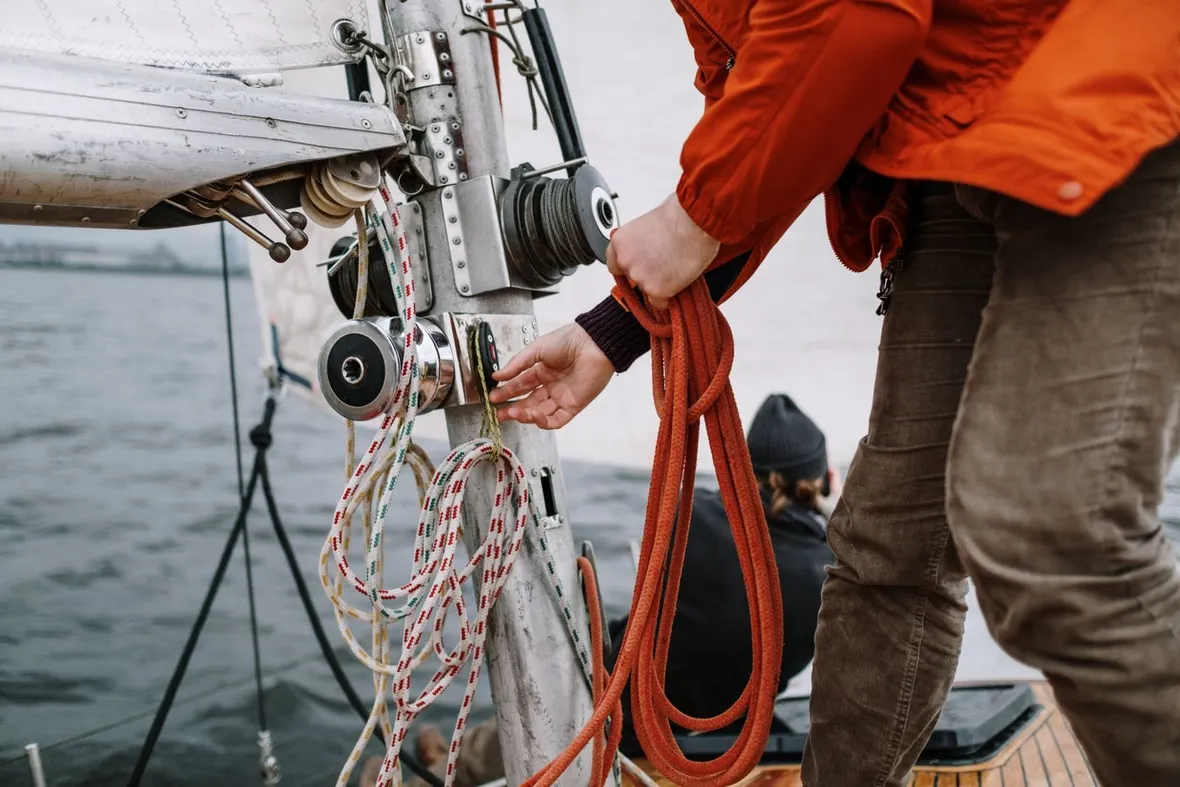 A crew member in an orange jacket holds a coiled rope at a yacht mast, the kind of sailing an RYA logbook records.