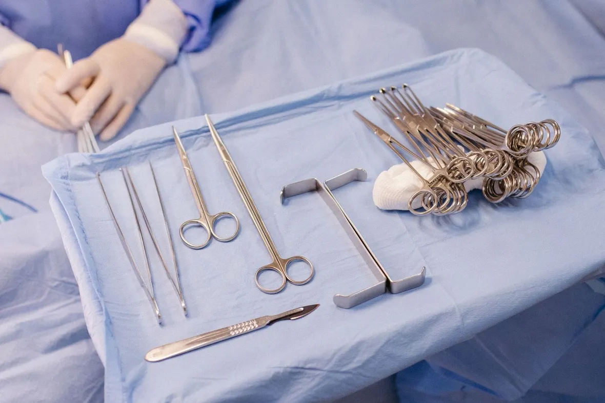 Surgical instruments laid out on a sterile blue drape with gloved hands working behind, the operating cases trainees record in a logbook.