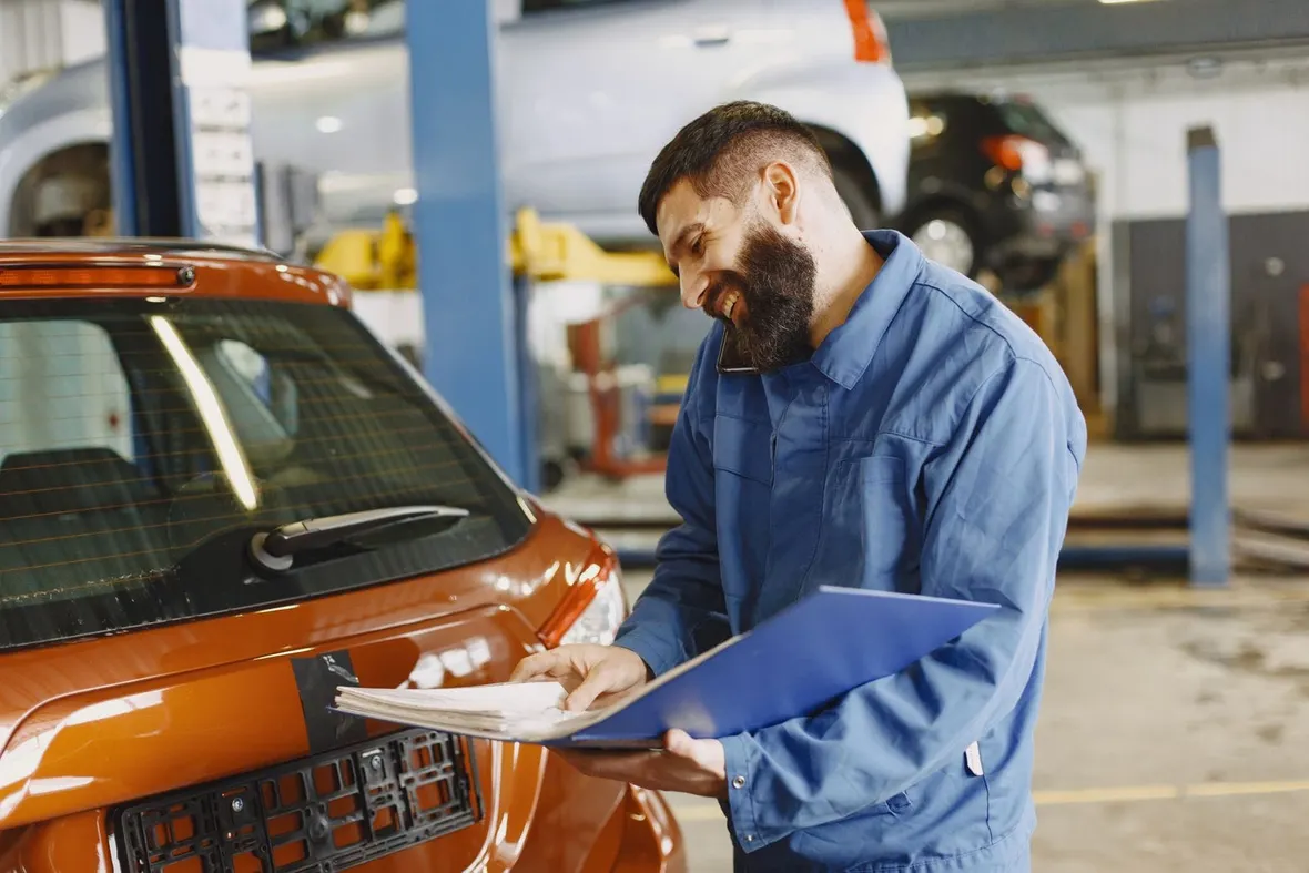 A mechanic in blue overalls checks papers in a folder beside a car in a garage, much like verifying a V5C logbook.